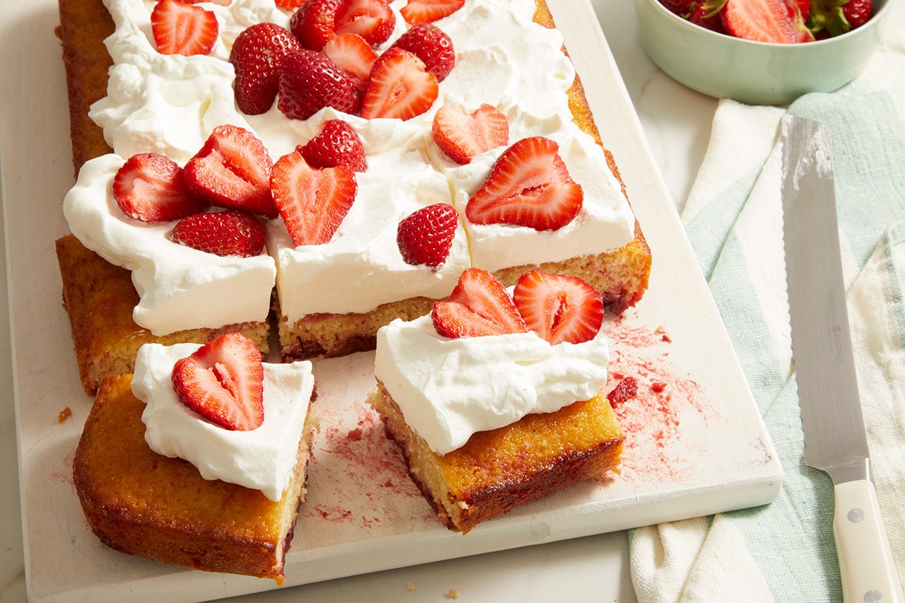 A sheet cake topped with whipped cream and sliced strawberries, with a few pieces cut and served on a white tray. A knife and a bowl of strawberries are nearby.