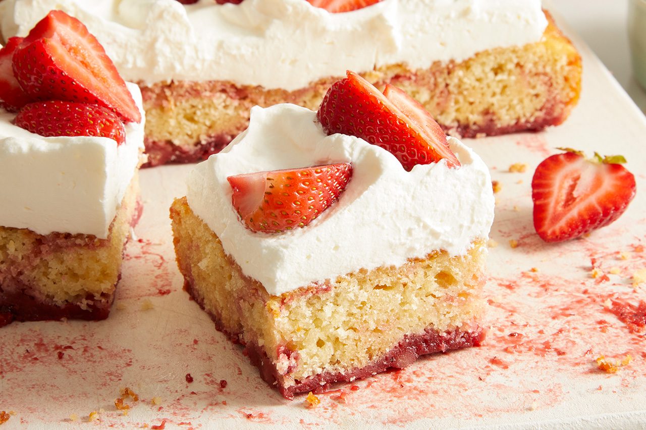 A square slice of cake topped with whipped cream and fresh strawberry pieces, with more cake slices and strawberries in the background on a white surface.