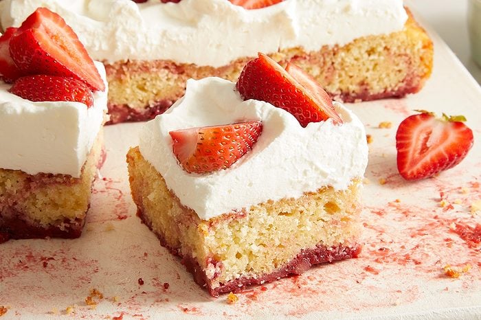 A square slice of cake topped with whipped cream and fresh strawberry pieces, with more cake slices and strawberries in the background on a white surface.