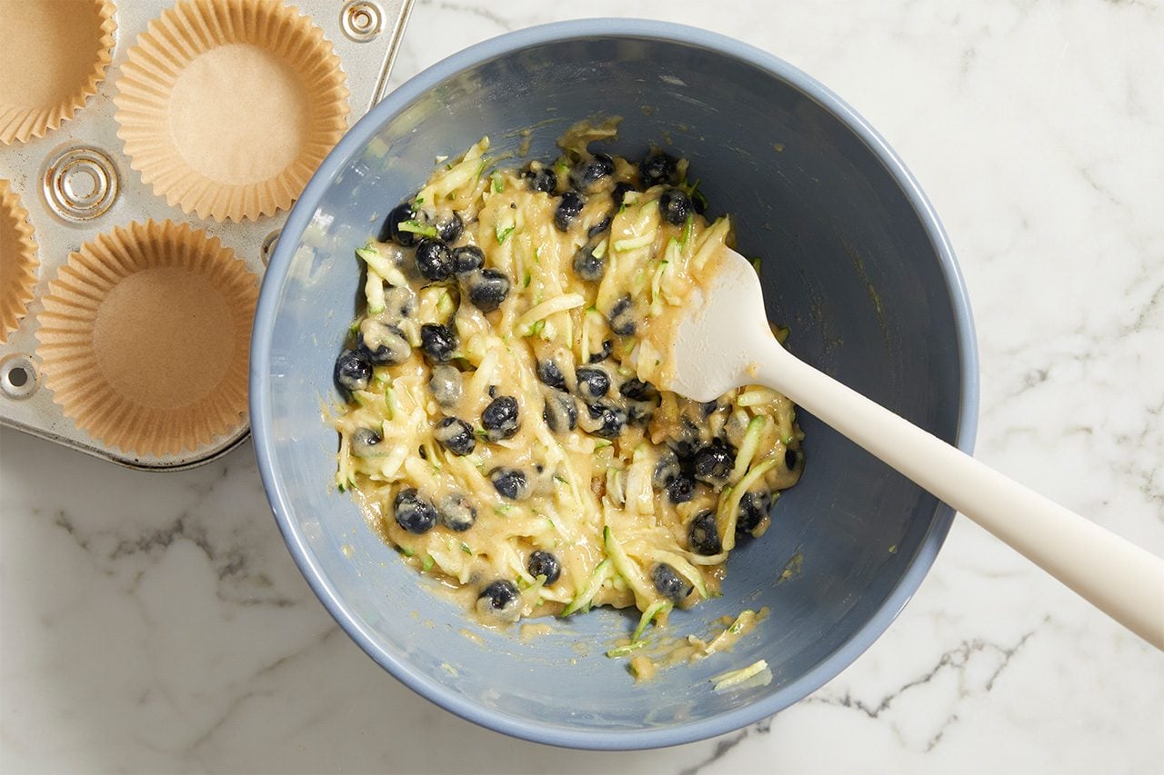 Overhead horizontal image of a light blue mixing bowl filled with a batter mixture. Shredded zucchini and whole blueberries are being stirred into the batter with a white spatula. A metal muffin tin with paper liners sits on a white marble countertop to the left.