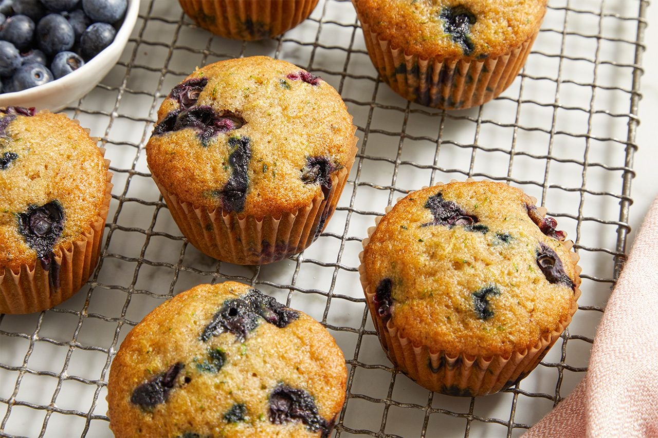 Overhead horizontal image of several freshly baked blueberry zucchini muffins resting on a silver wire cooling rack. A white bowl filled with fresh blueberries is partially visible in the upper left background, and a pink napkin is in the lower right corner.