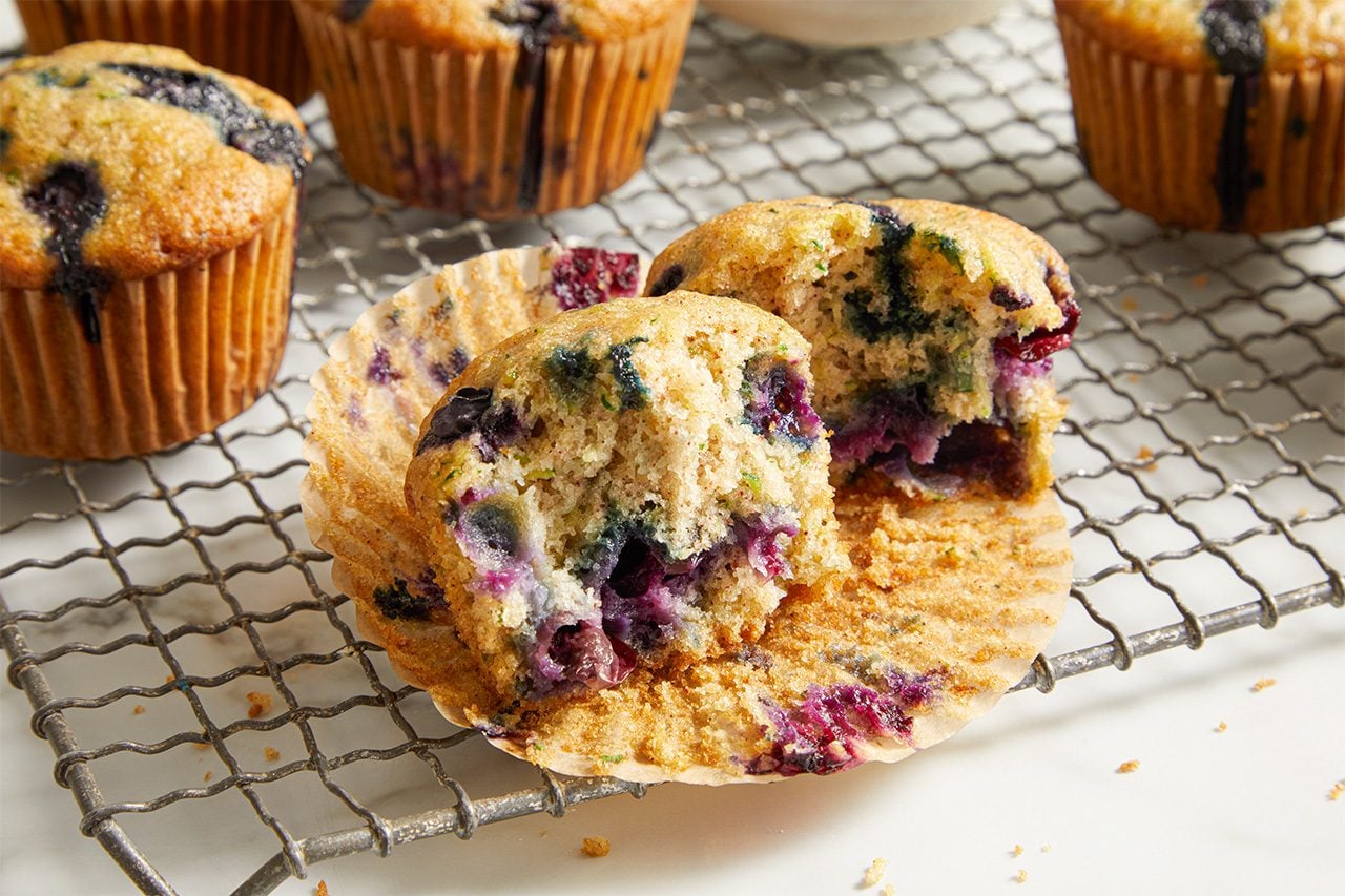 Eye Level horizontal image of a blueberry zucchini muffin torn in half, showcasing the moist interior with bursts of blueberries and flecks of green zucchini. Several other whole blueberry zucchini muffins are visible in the background, all resting on a metal cooling rack.