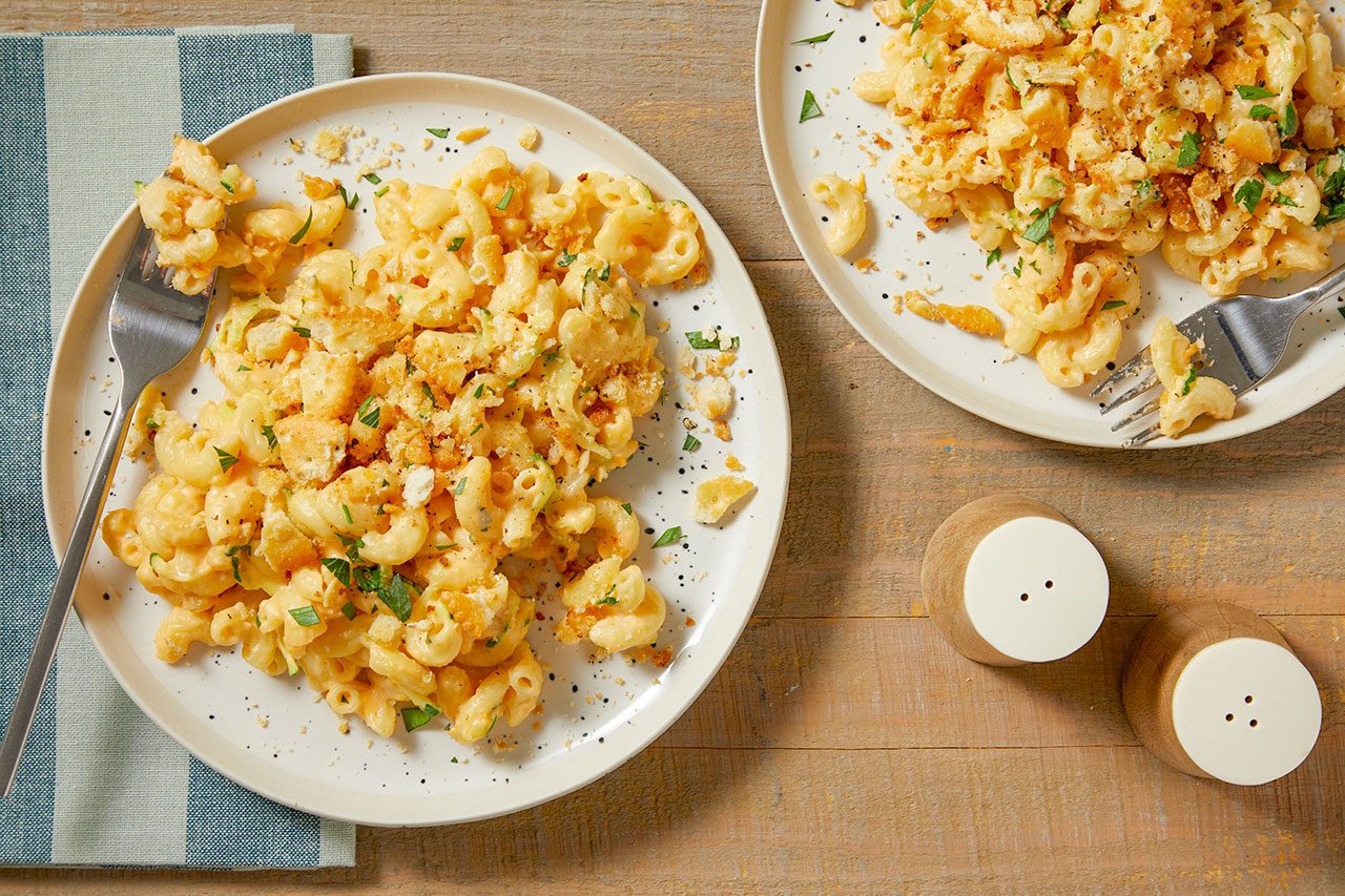 Overhead horizontal image of two servings of macaroni and cheese, topped with toasted breadcrumbs and fresh herbs. One plate features a fork, and both are placed on a wooden surface alongside a striped napkin and salt and pepper shakers.