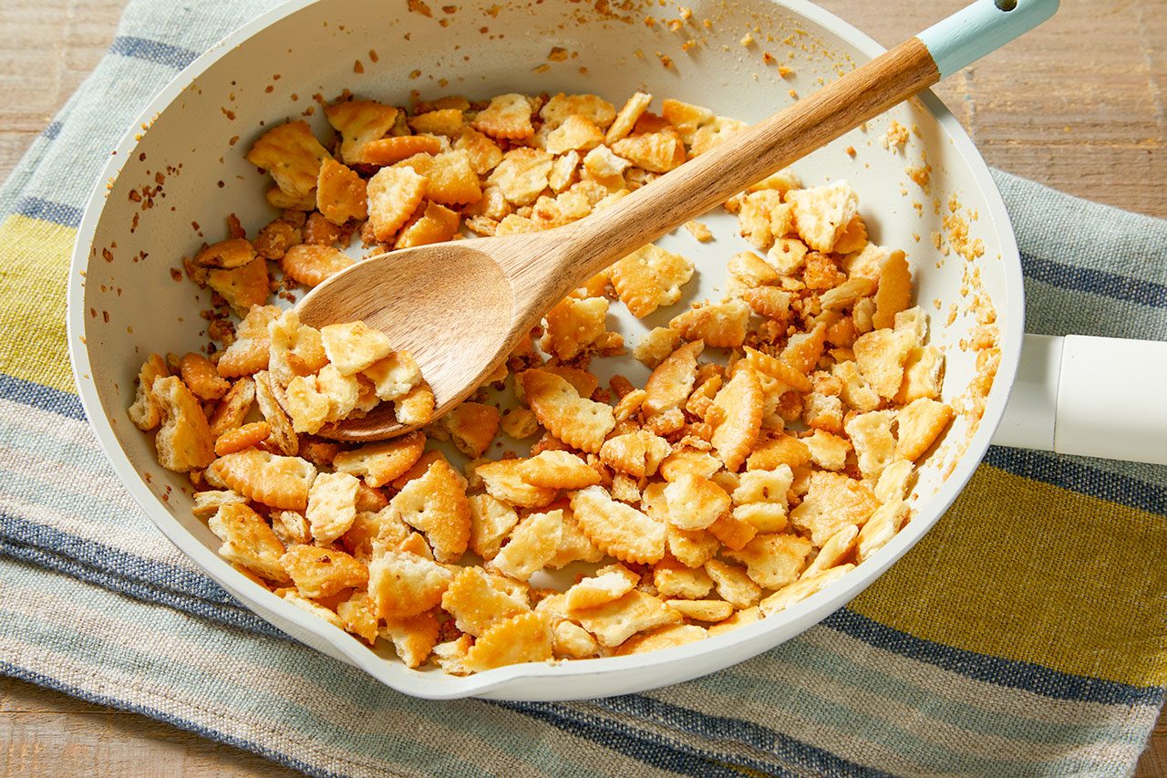 Overhead horizontal image of a white pan filled with toasted, broken crackers. A wooden spoon with a light blue handle rests in the pan, mixing the crackers that are lightly browned.