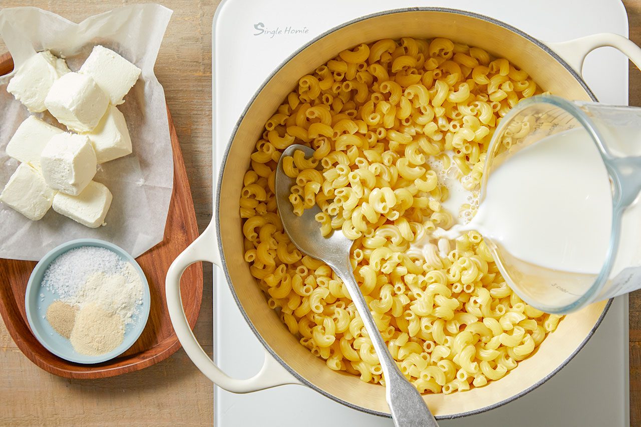 Overhead horizontal image showing milk being poured from a measuring cup into a white pot filled with cooked elbow macaroni. To the left, cubes of cream cheese rest on parchment paper on a wooden board, with a small dish containing salt, garlic powder, and onion powder.