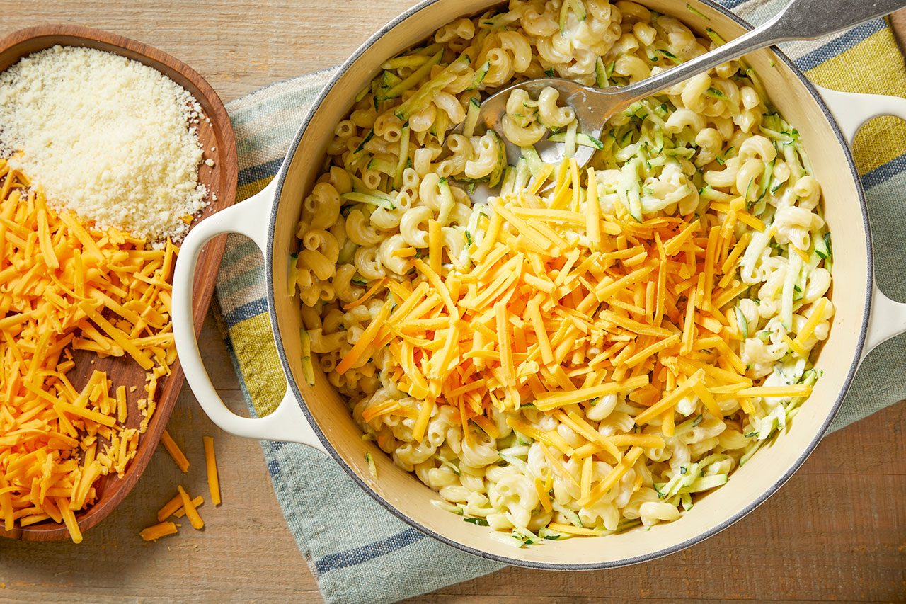 Overhead horizontal image of a white enamel Dutch oven filled with creamy macaroni pasta and shredded zucchini. A generous pile of shredded cheddar cheese has been added to one side of the pasta, with a serving spoon visible within the pot. A wooden tray alongside the pot holds additional shredded cheddar cheese and grated Parmesan cheese.