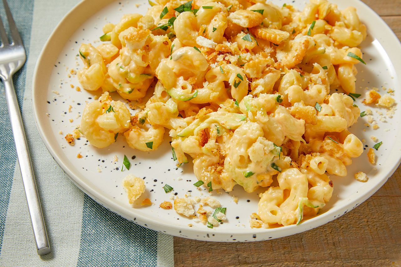 Eye Level horizontal image of creamy macaroni and cheese mixed with green zucchini noodles, topped with golden-brown breadcrumbs and fresh green herbs. The dish is served on an off-white plate with black speckles, with a fork resting on a striped blue and white linen beside it.