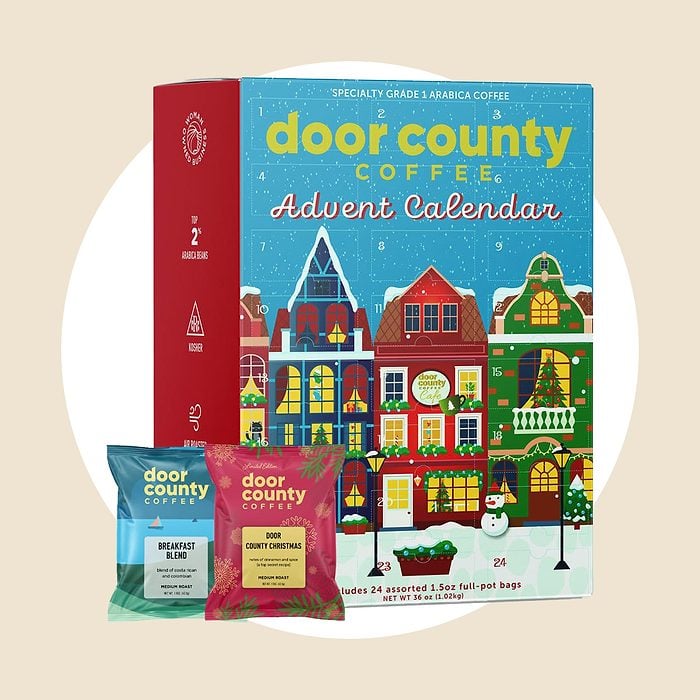 A Door County Coffee Advent Calendar box with a colorful holiday village design, alongside two sample coffee packets labeled “Breakfast Blend” and “Dark Voyage.”.