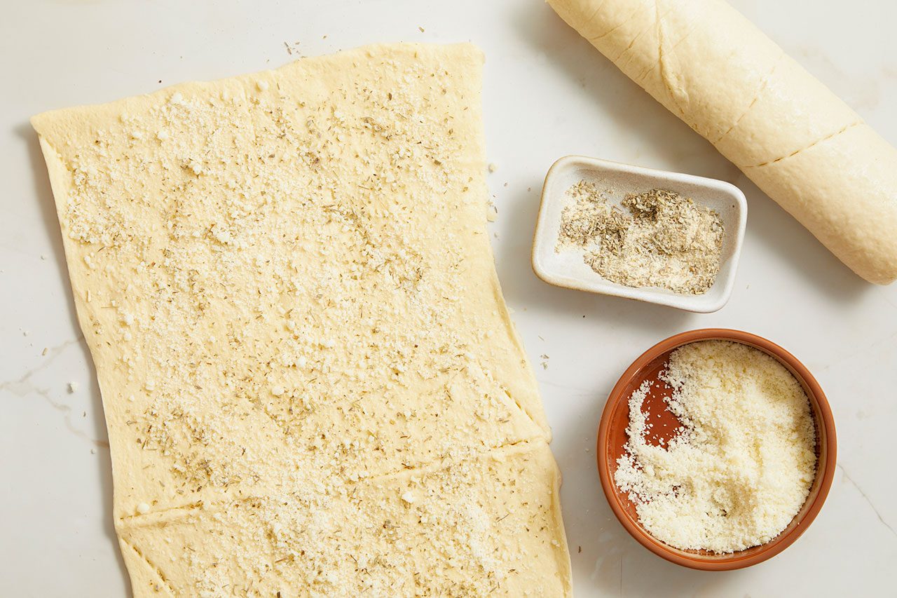 Overhead horizontal image of a sheet of raw dough sprinkled with grated Parmesan cheese and dried herbs. A bowl of grated Parmesan cheese and a small dish containing dried herbs and spices are nearby, along with a log of raw dough.