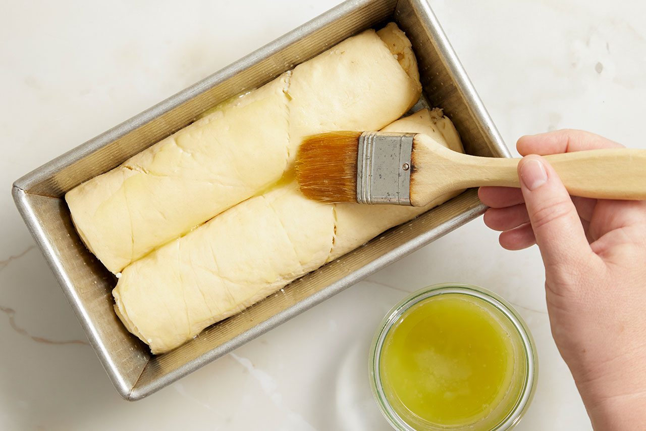 Overhead horizontal image of a hand using a pastry brush to coat two rolls of uncooked dough with melted butter or oil. The dough is placed inside a silver loaf pan, with a small glass bowl of the yellow liquid visible to the right.