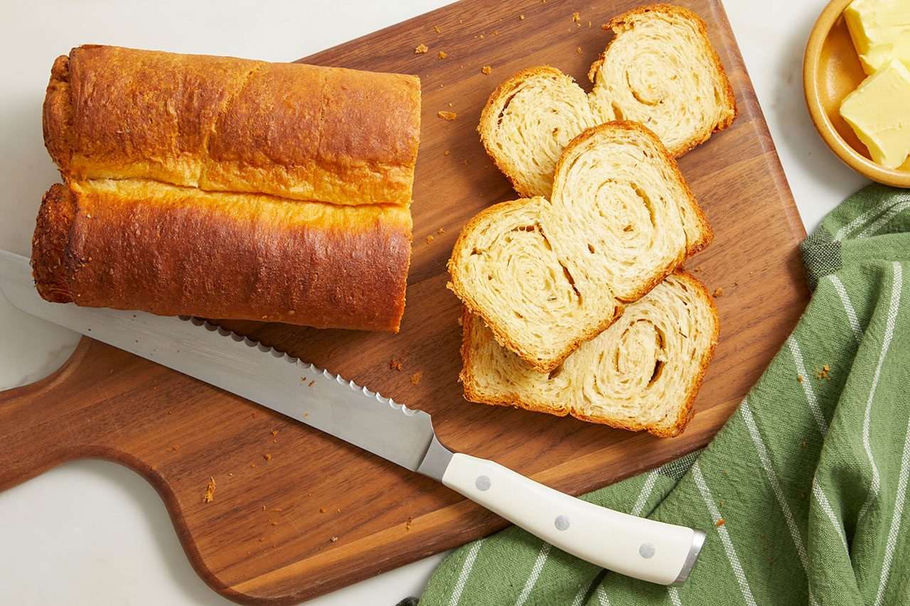 Overhead horizontal image of a golden brown loaf of bread, partially sliced, resting on a wooden cutting board. Several slices of bread with a swirled interior are arranged next to a serrated knife. Pats of butter are visible in a small wooden bowl in the background.