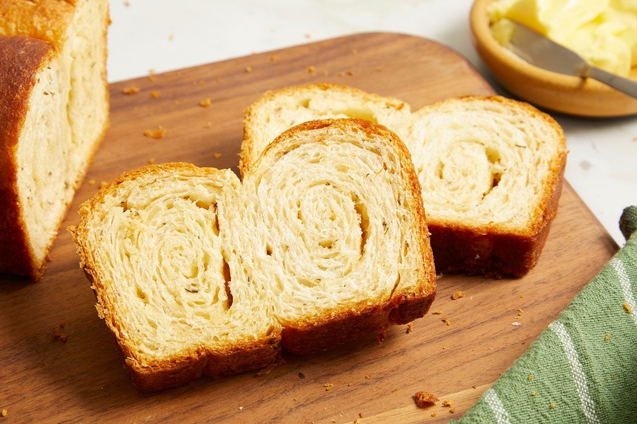 Eye Level horizontal image of freshly baked, sliced bread on a wooden cutting board. Several thick slices are fanned out next to a partial loaf, revealing their swirled interior texture. In the background, a small wooden bowl holds pats of butter and a butter knife, with a green striped towel visible in the foreground.