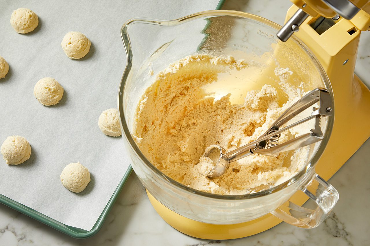 Overhead horizontal image of cookie dough being scooped onto a parchment-lined baking sheet. A glass mixing bowl filled with dough and a metal cookie scoop are next to the sheet, with a yellow stand mixer partially visible.