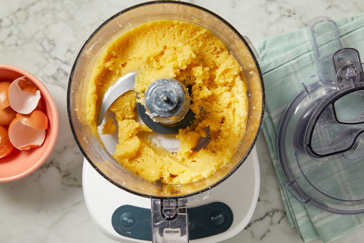 Overhead horizontal image of a food processor containing a bright yellow dough mixture. Next to it, a pink bowl holds cracked eggshells, and the food processor lid rests on a light blue kitchen towel on a marble countertop.