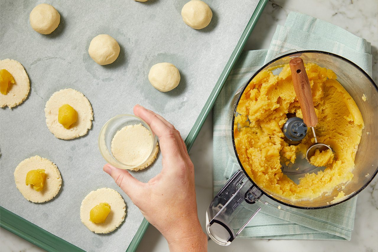 Overhead horizontal image of a hand holding a small bowl containing cookie dough, while preparing unbaked cookies on a parchment-lined baking sheet. Several light-colored cookie dough balls are visible, along with flattened discs of dough containing a yellow fruit filling. A food processor filled with the yellow filling and a small scooper sits to the right.