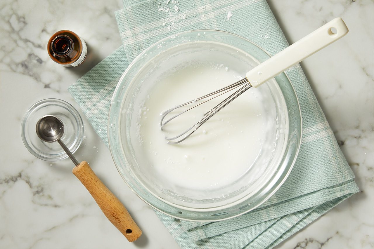 Overhead horizontal image of a whisk resting in a clear glass bowl filled with white icing or glaze. To the left, a brown bottle of extract and a measuring spoon in a small glass bowl with clear liquid are visible on a marble countertop, alongside a light blue-green kitchen towel with scattered powdered sugar.