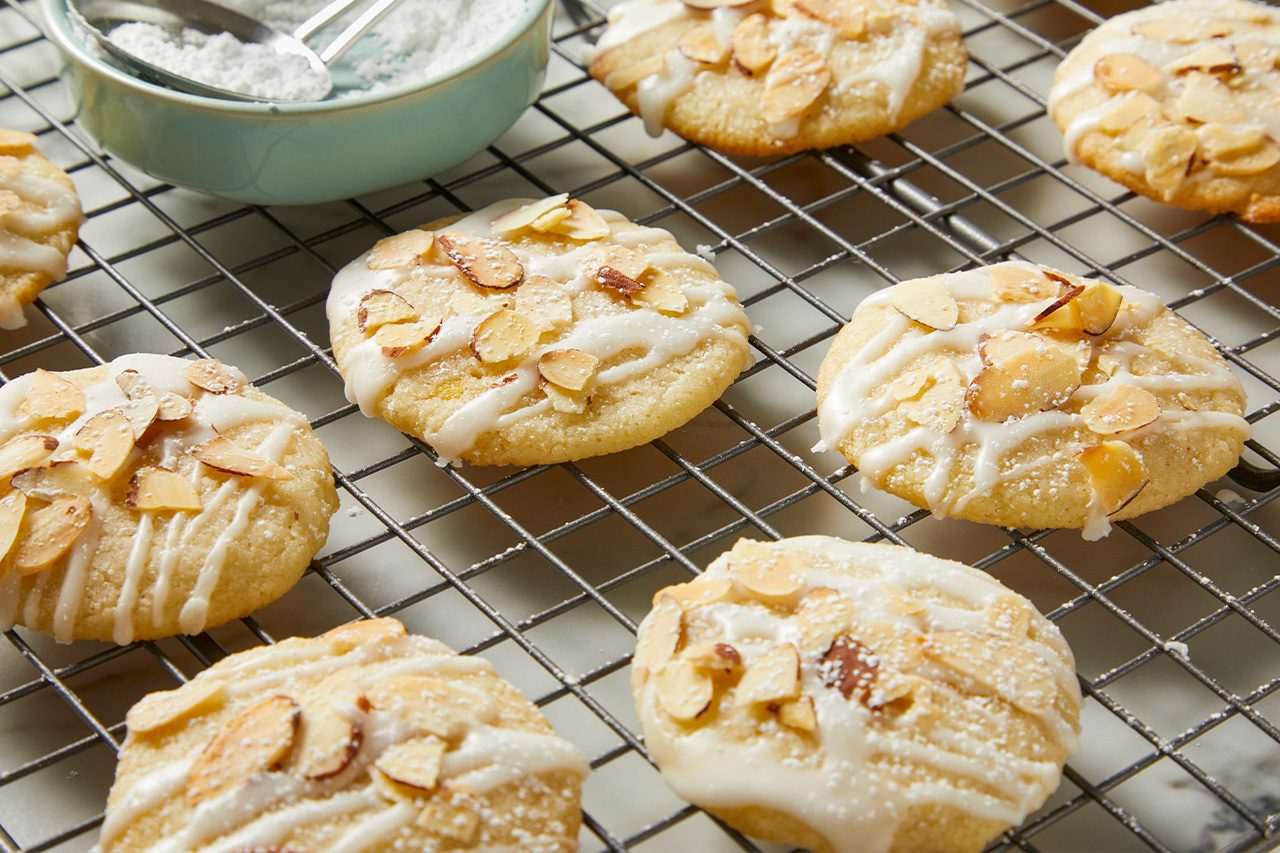 Overhead horizontal image of several glazed cookies topped with sliced almonds, cooling on a wire rack. A light blue bowl of powdered sugar with a silver spoon rests behind the cookies, some of which are dusted with additional powdered sugar.