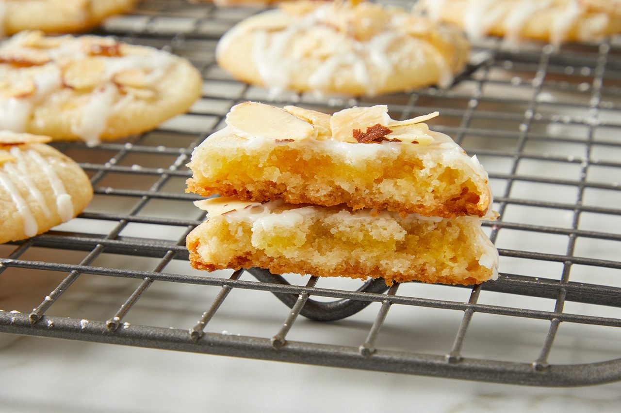Eye Level horizontal image of a cookie broken in half, stacked on itself, revealing its soft, yellowish interior. The cookie is topped with a white glaze and slivered almonds, sitting on a dark grey cooling rack with other cookies blurred in the background.