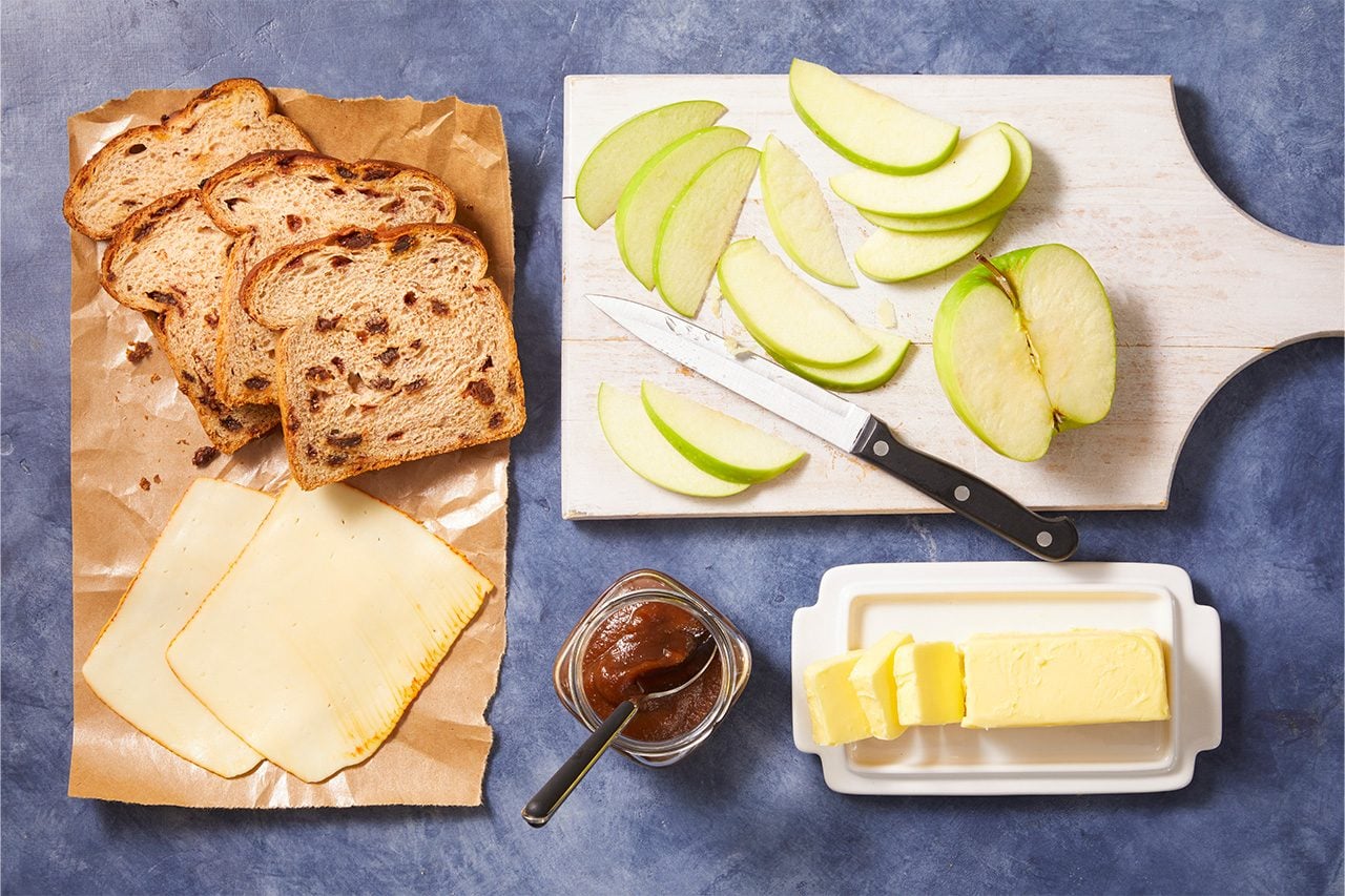 Overhead horizontal image of ingredients arranged on a blue surface. Slices of cinnamon raisin bread and deli cheese are placed on brown paper, while sliced green apples and a knife rest on a white cutting board. A jar of apple butter with a spoon and a partially sliced stick of butter in a white dish are also visible.