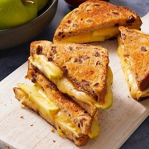 Eye Level vertical image of a golden brown grilled cheese sandwich, made with raisin bread, stacked on a white cutting board. The sandwich is cut in half, revealing melted cheese, green apple slices, and a rich, brown spread. Whole green apples in a bowl and a jar of the brown spread are visible in the background.