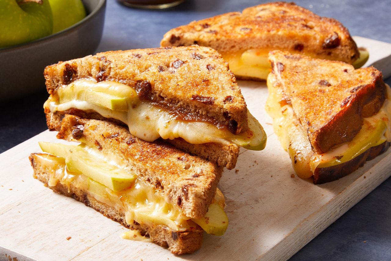 Eye Level horizontal image of three grilled cheese sandwiches made with cinnamon raisin bread, melted cheese, and slices of green apple. The golden-brown sandwiches are presented on a white wooden cutting board, with a bowl of green apples visible in the soft-focus background.