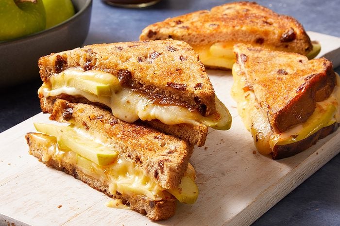 Eye Level horizontal image of three grilled cheese sandwiches made with cinnamon raisin bread, melted cheese, and slices of green apple. The golden-brown sandwiches are presented on a white wooden cutting board, with a bowl of green apples visible in the soft-focus background.
