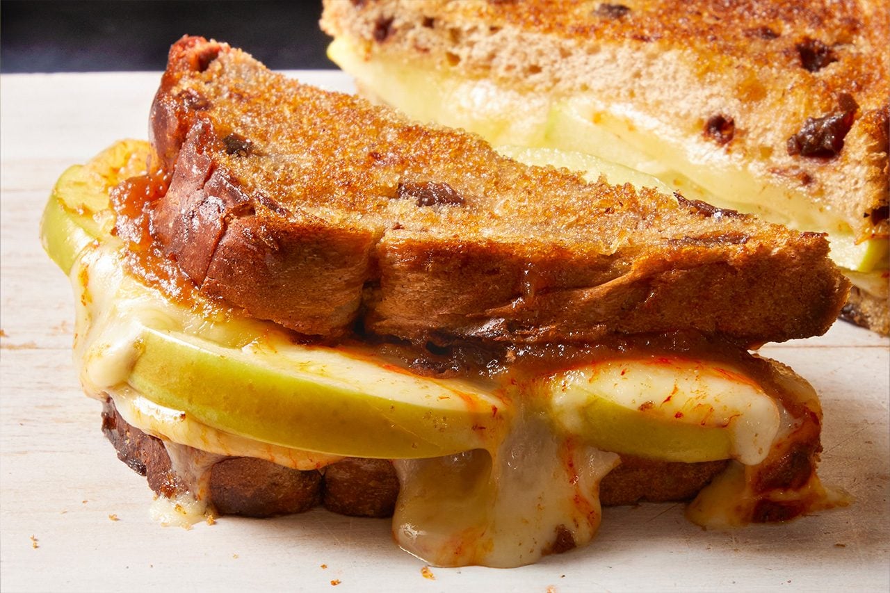 Eye Level horizontal image of a grilled cheese sandwich cut in half, revealing its delicious cross-section. This sandwich is made with toasted cinnamon raisin bread, melted cheese, and slices of green apple. Fruit preserves are visible along with the oozing cheese.