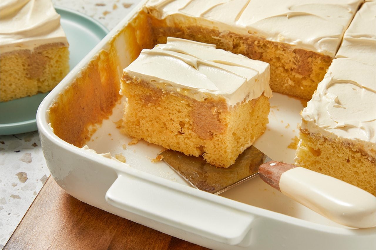 Eye Level horizontal image of a slice of sheet cake being lifted from a white rectangular baking dish with a cake server. The yellow cake has a creamy white frosting on top and a brown caramel or cinnamon swirl visible within the cake layers.