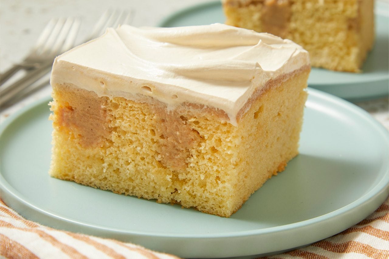 Eye Level horizontal image of a slice of cake on a light blue plate. The cake features a light yellow sponge base, a middle layer of light brown filling or frosting, and is topped with swirls of white piped frosting. A striped cloth is visible in the foreground, and blurred forks are in the background.
