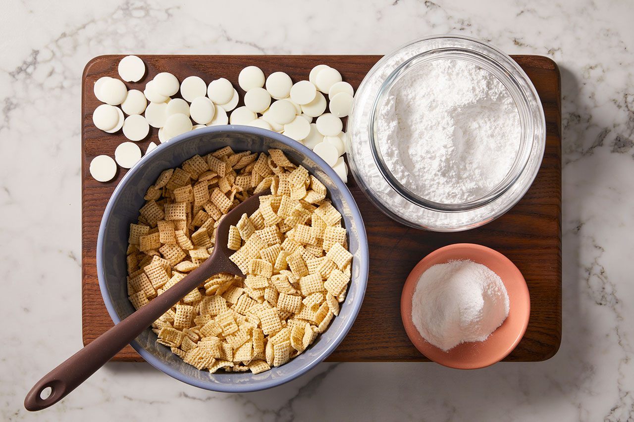 Overhead horizontal image of ingredients for a recipe, including a blue bowl of square-shaped cereal with a brown silicone spoon, a pile of white chocolate melting wafers, a glass jar of powdered sugar, and a small dish of granulated sugar. These items are arranged on a wooden cutting board on a marble countertop.
