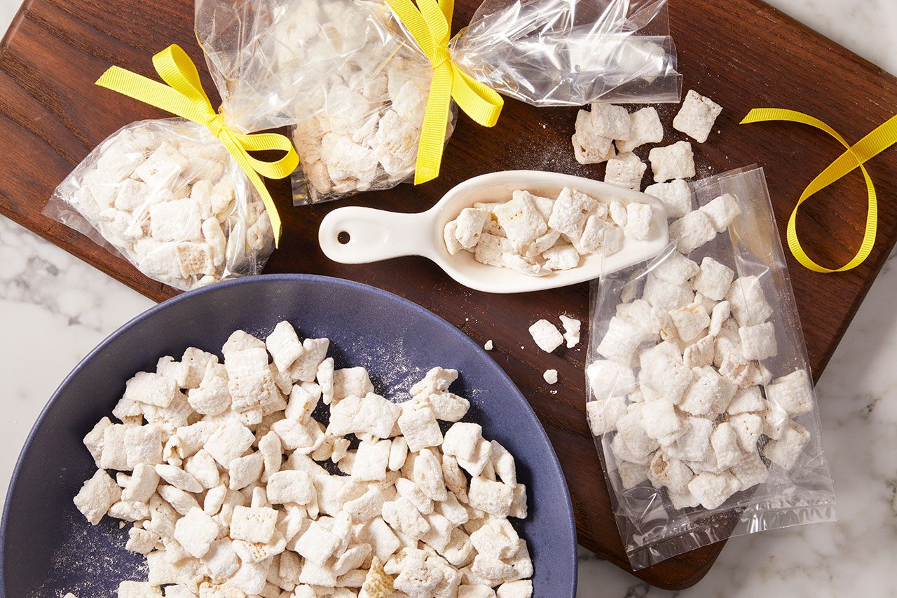 Overhead horizontal image of a sweet cereal snack being packaged. A blue bowl is filled with the powdered sugar-coated snack, with some also in a white scoop and in several clear plastic bags, two of which are tied with bright yellow ribbons. The items are arranged on a wooden cutting board against a marble background.