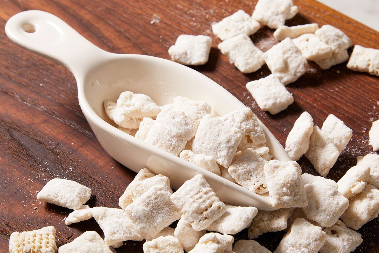 Eye level horizontal image of white, square-shaped snack pieces, heavily coated in powdered sugar, overflowing from a white ceramic scoop. More pieces are scattered across a dark wooden background, showcasing a crunchy, sweet treat.