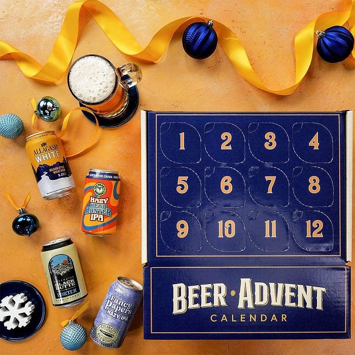 A Beer Advent Calendar box with numbered doors sits on a yellow surface, surrounded by beer cans, a glass of beer, blue ornaments, and a yellow ribbon.