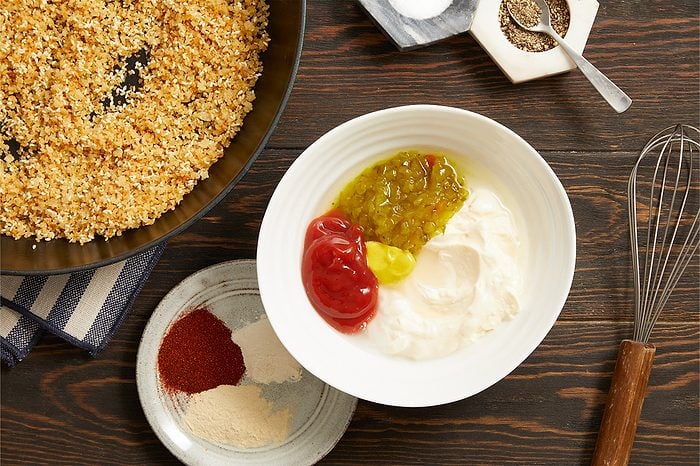 Overhead horizontal image of a white bowl containing mayonnaise, ketchup, mustard, and green relish. On a dark wooden surface, a small plate holds red paprika, white garlic powder, and light brown onion powder, alongside a pan of toasted breadcrumbs with sesame seeds. Salt, pepper, and a whisk are also visible on the rustic background.
