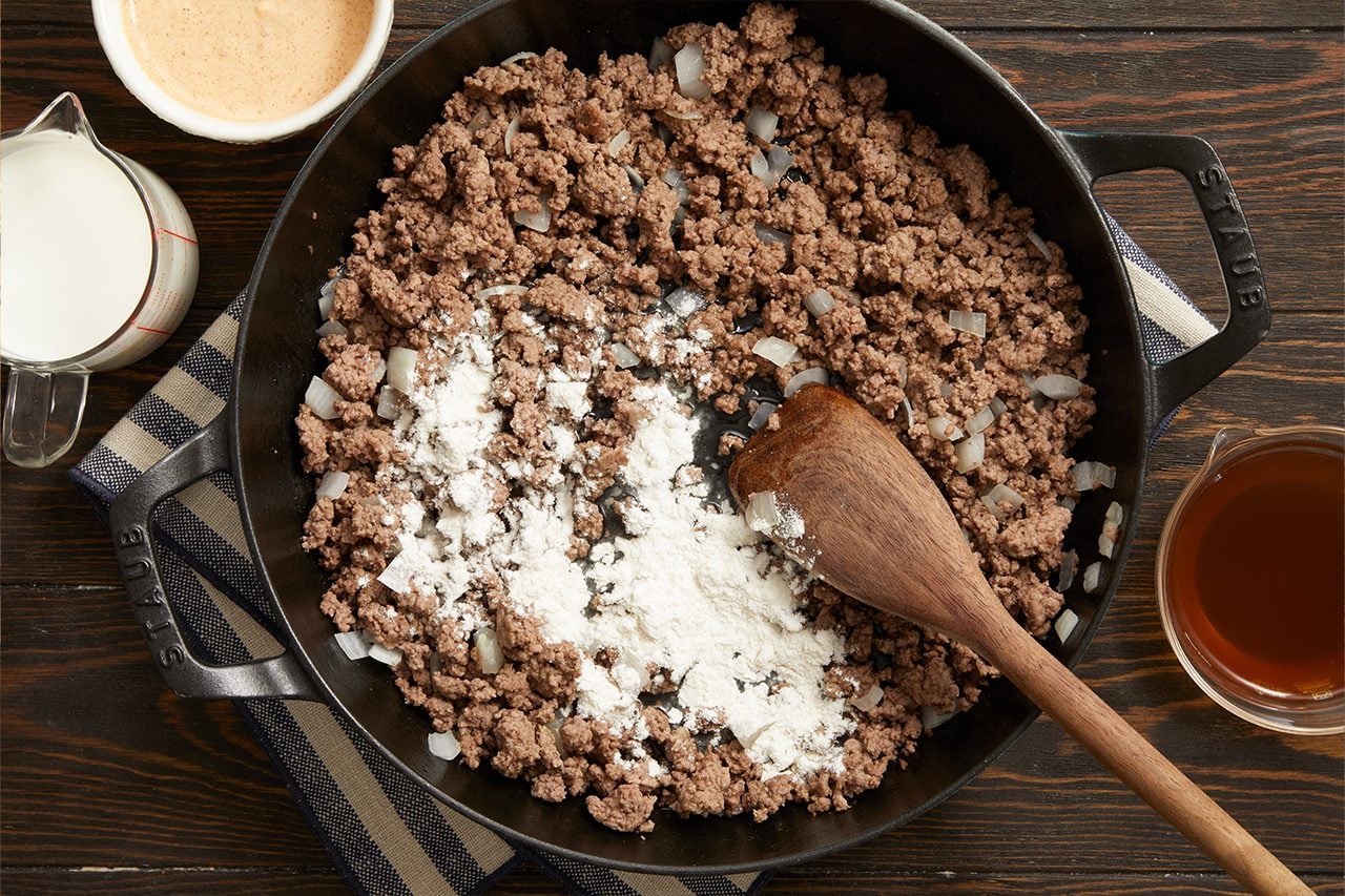 Overhead horizontal image of cooked ground beef and chopped onions in a black skillet. A pile of white flour rests on the meat, next to a wooden spoon. Nearby are a measuring cup of milk, a bowl of light brown sauce, and a measuring cup of dark liquid.