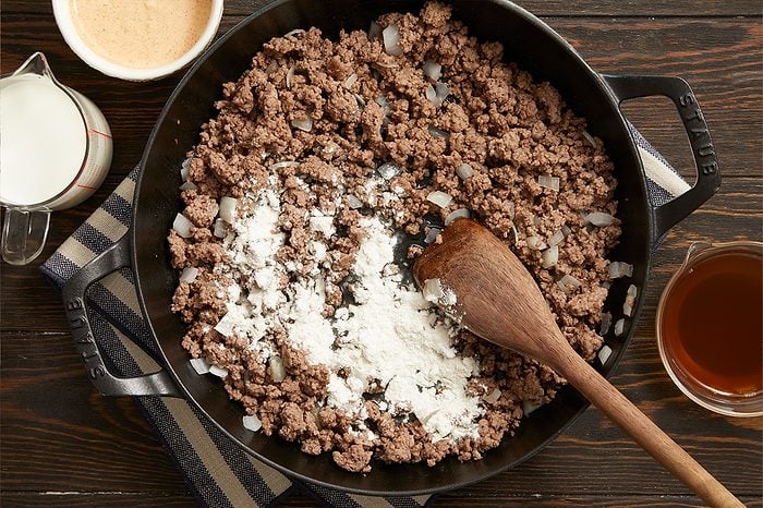 Overhead horizontal image of cooked ground beef and chopped onions in a black skillet. A pile of white flour rests on the meat, next to a wooden spoon. Nearby are a measuring cup of milk, a bowl of light brown sauce, and a measuring cup of dark liquid.