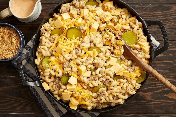 Overhead horizontal image of a cast-iron skillet filled with a macaroni dish. The skillet contains elbow macaroni mixed with ground meat, topped with shredded cheddar cheese, cubed American cheese, and dill pickle slices, with a wooden spoon resting in the dish. A small pitcher of sauce and a bowl of breadcrumbs are visible on the dark wooden surface beside the skillet.