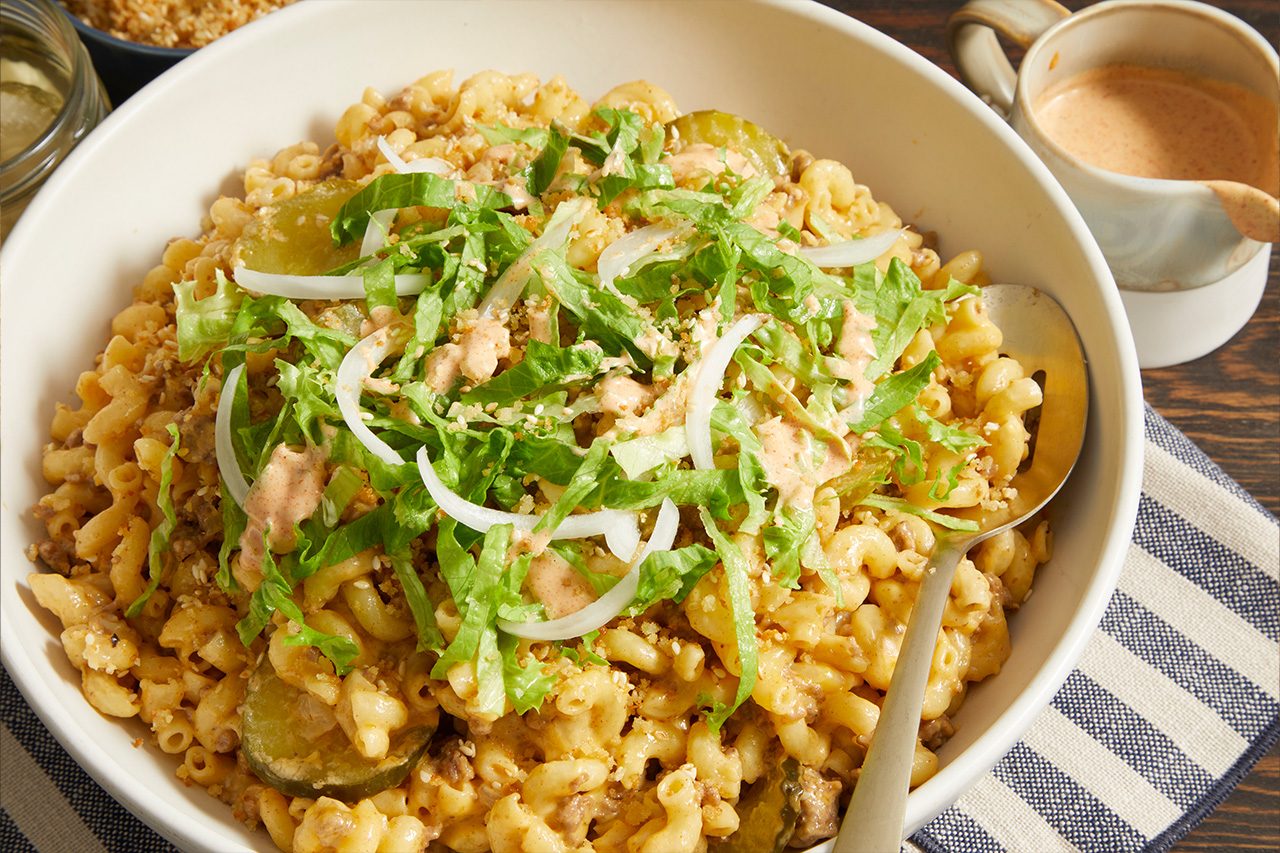Overhead horizontal image of a large white bowl filled with macaroni and ground beef in a creamy cheese sauce. The dish is topped with shredded lettuce, thin slices of white onion, pickles, a pinkish burger-style sauce, and sesame seeds, with a serving spoon resting in the bowl.