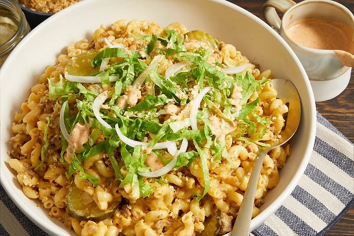 Overhead horizontal image of a large white bowl filled with macaroni and ground beef in a creamy cheese sauce. The dish is topped with shredded lettuce, thin slices of white onion, pickles, a pinkish burger-style sauce, and sesame seeds, with a serving spoon resting in the bowl.