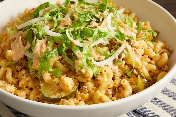 Eye level horizontal image of a large white bowl filled with a mac and cheese dish. It is generously topped with shredded lettuce, sliced white onions, a creamy special sauce, and sesame seeds. Visible within the elbow macaroni are pieces of ground meat and sliced pickles.