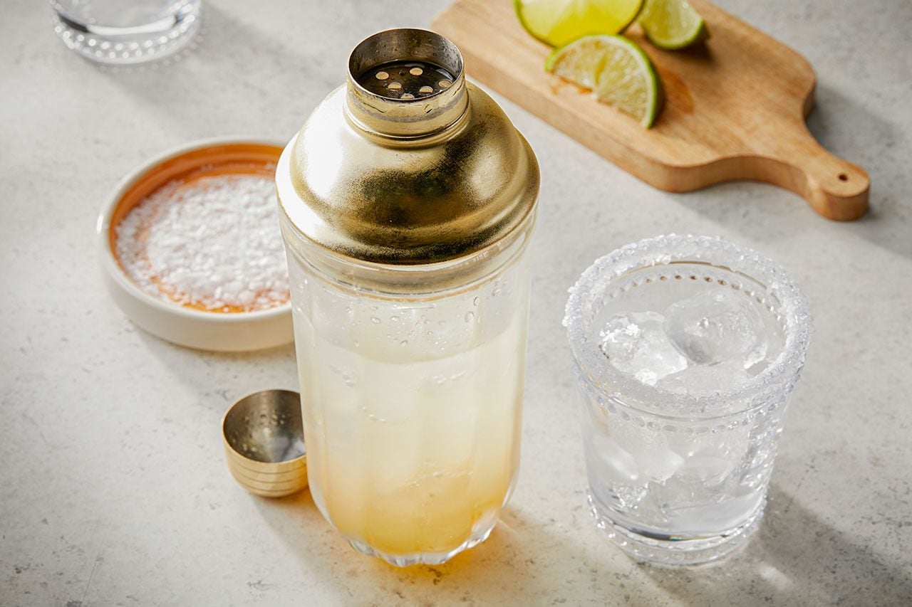 Eye Level horizontal image of a gold and clear cocktail shaker filled with a light-colored drink. Beside it, a glass rimmed with salt is full of ice, and a small dish holds additional salt. In the background, lime wedges rest on a wooden cutting board, suggesting ingredients for a refreshing beverage.