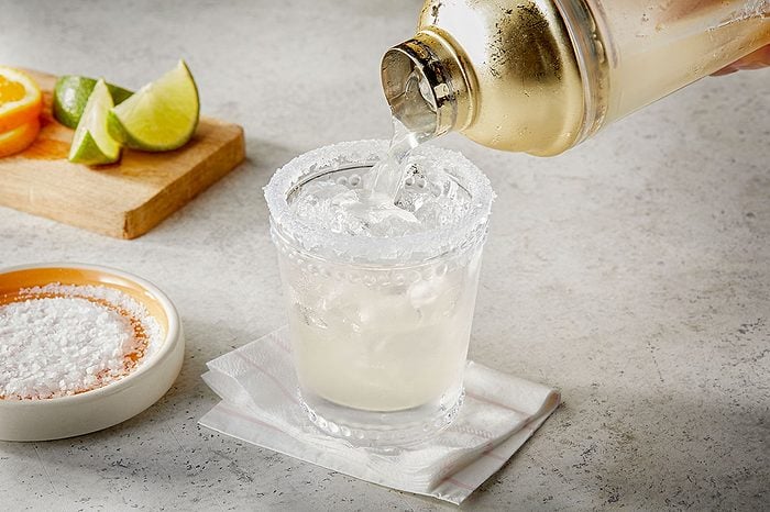 Eye Level horizontal image of a clear beverage being poured from a gold cocktail shaker into a salt-rimmed glass filled with crushed ice. In the background, lime wedges and orange slices rest on a wooden cutting board, and a small bowl of flaky salt is visible.
