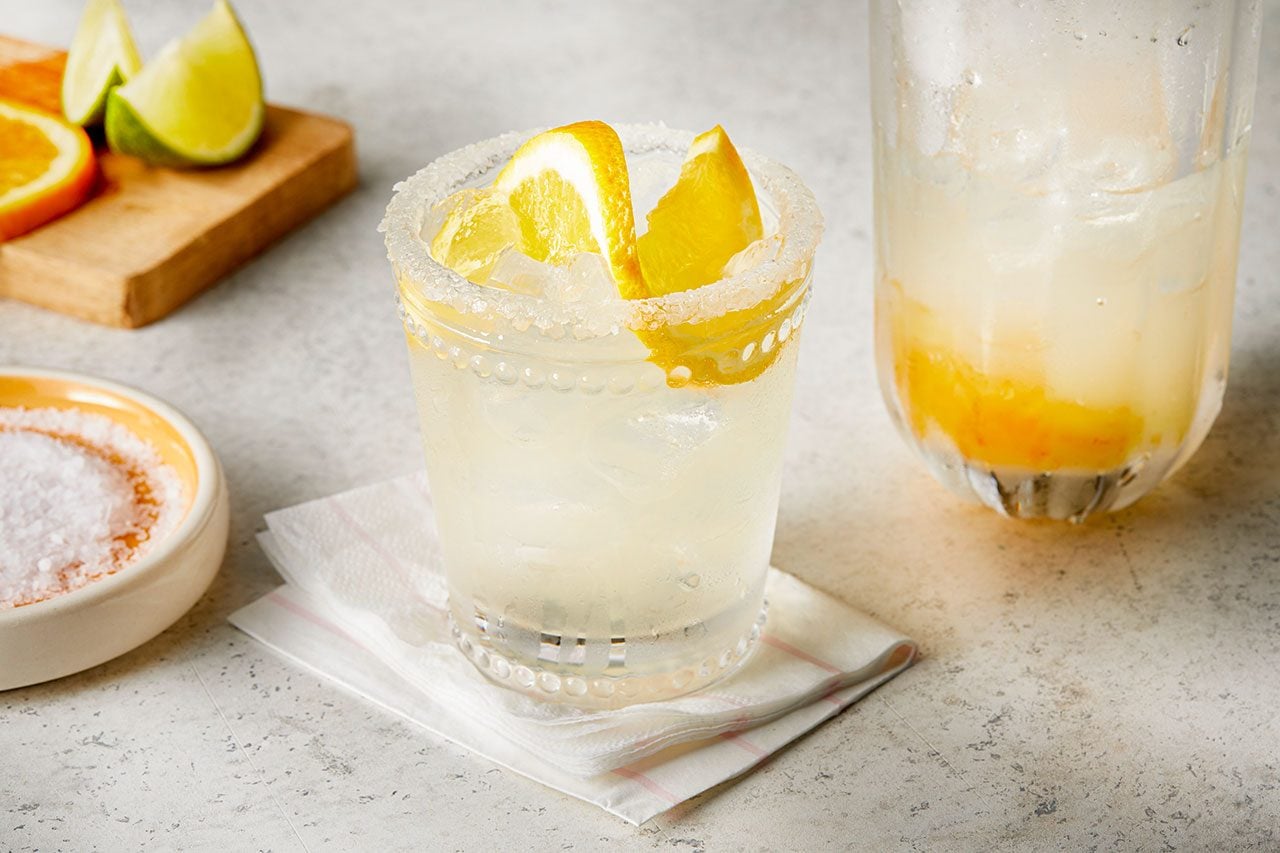 Eye Level horizontal image of a salt-rimmed glass filled with an iced beverage and garnished with lemon slices. A bowl of salt, orange, and lime wedges are visible in the background, along with another glass containing muddled fruit.