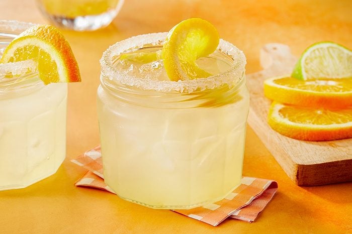 Eye Level horizontal image of two chilled, light yellow drinks served in hexagonal glasses with sugar-rimmed edges. The foreground drink is garnished with a lemon peel and contains ice, while the background drink has an orange slice on its rim. Sliced oranges and a lime wedge rest on a wooden board to the right.