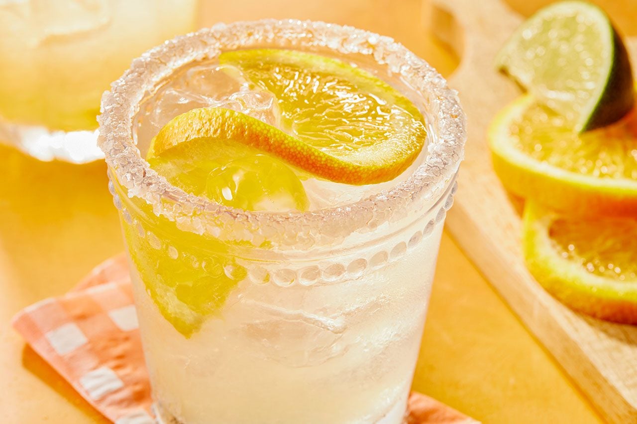 Eye-level horizontal image of a glass filled with ice and a beverage, garnished with lemon slices and a salted rim. In the background, additional lemon slices and a lime wedge rest on a wooden cutting board next to a checkered napkin. 