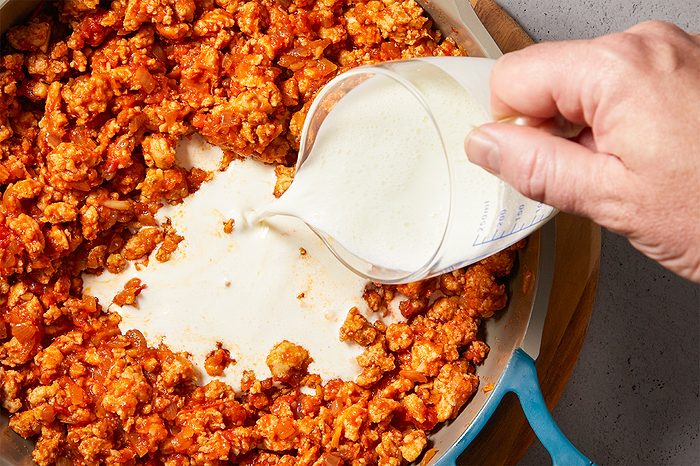 Overhead horizontal image of a hand pouring milk or cream from a measuring cup into a blue pan. The pan contains cooked ground meat, likely ground chicken or turkey, mixed with a reddish sauce and diced onions.