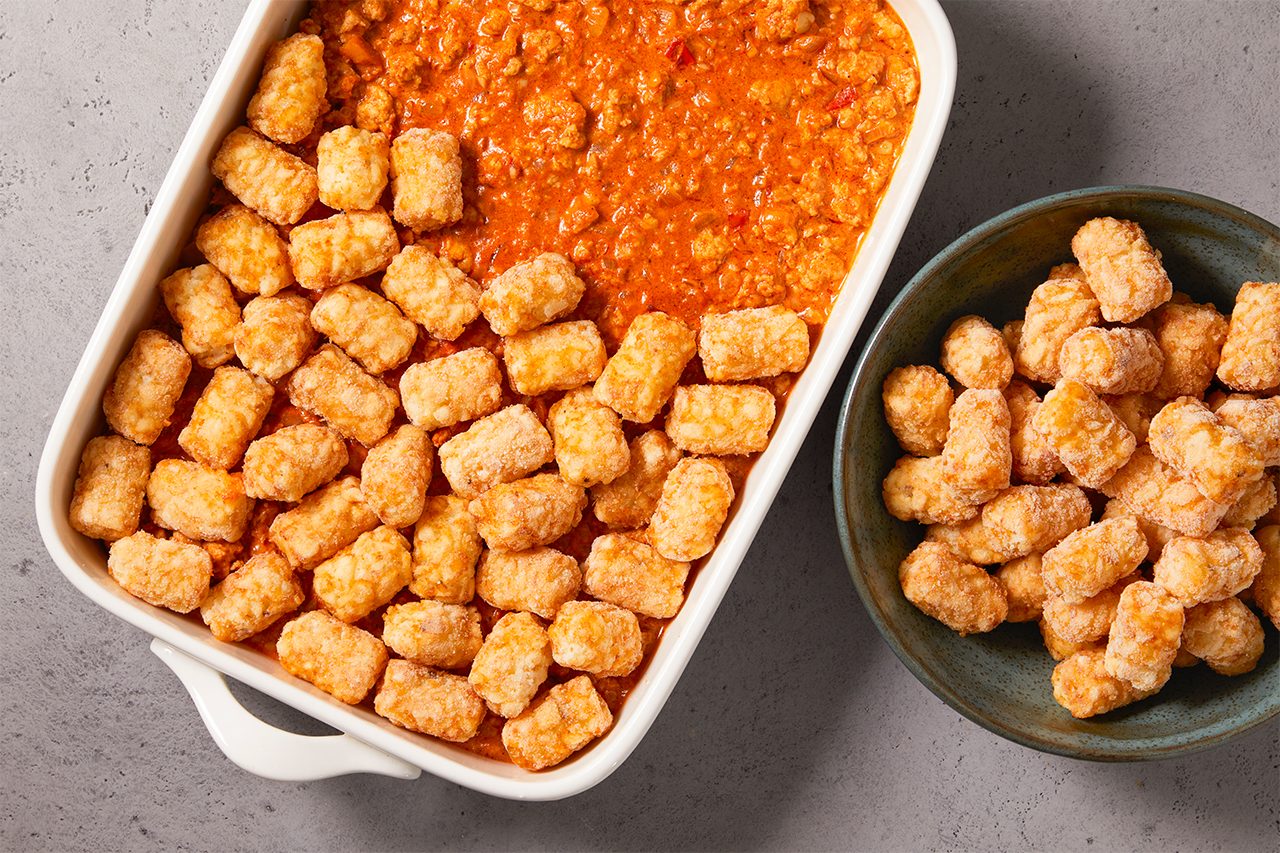 Overhead horizontal image of a white baking dish containing a ground meat and sauce mixture, partially topped with uncooked tater tots. A separate blue bowl filled with additional uncooked tater tots is visible to the right, ready to be added to the dish.
