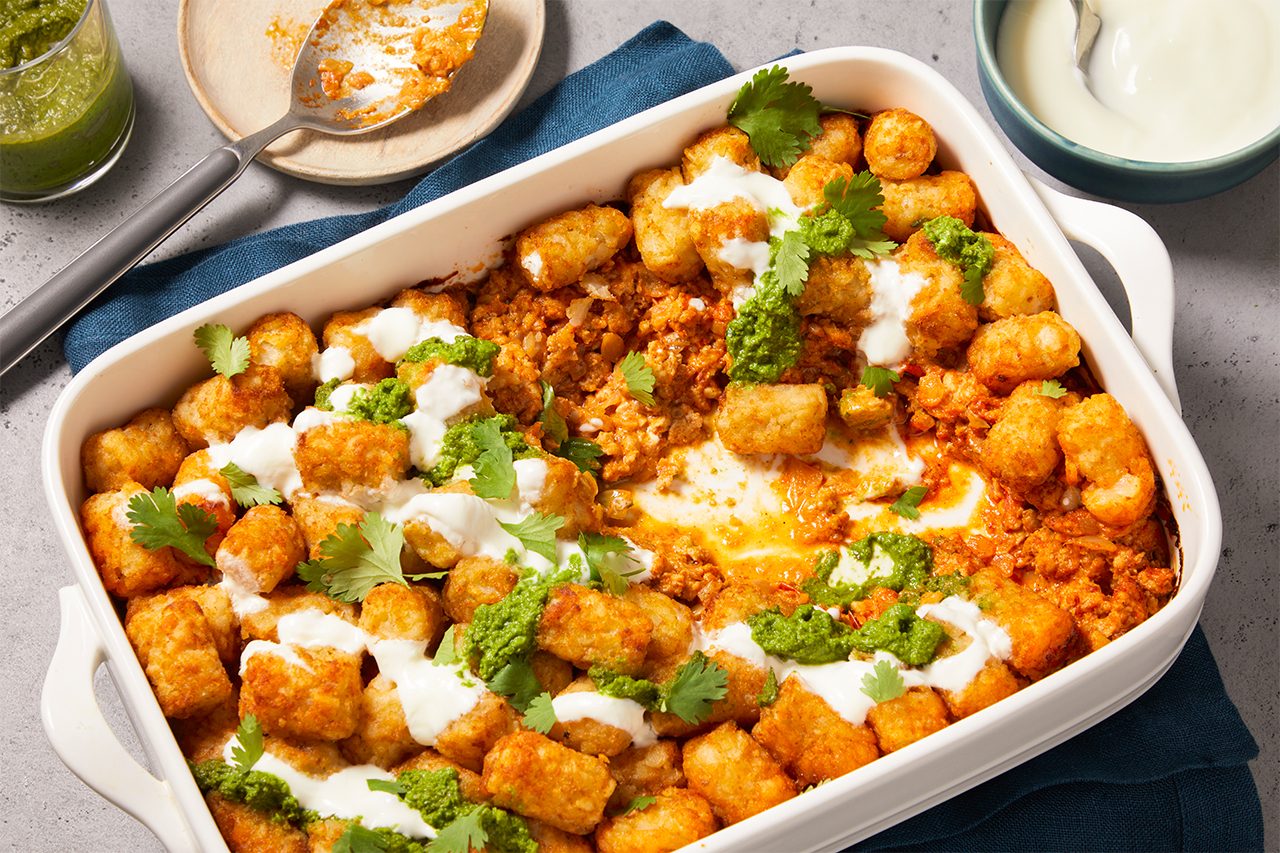 Overhead horizontal image of a tater tot casserole in a white baking dish, garnished with dollops of white cream, green pesto, and fresh cilantro. A spoon with casserole mixture on a small plate and a bowl of white cream are placed beside the dish on a blue napkin.