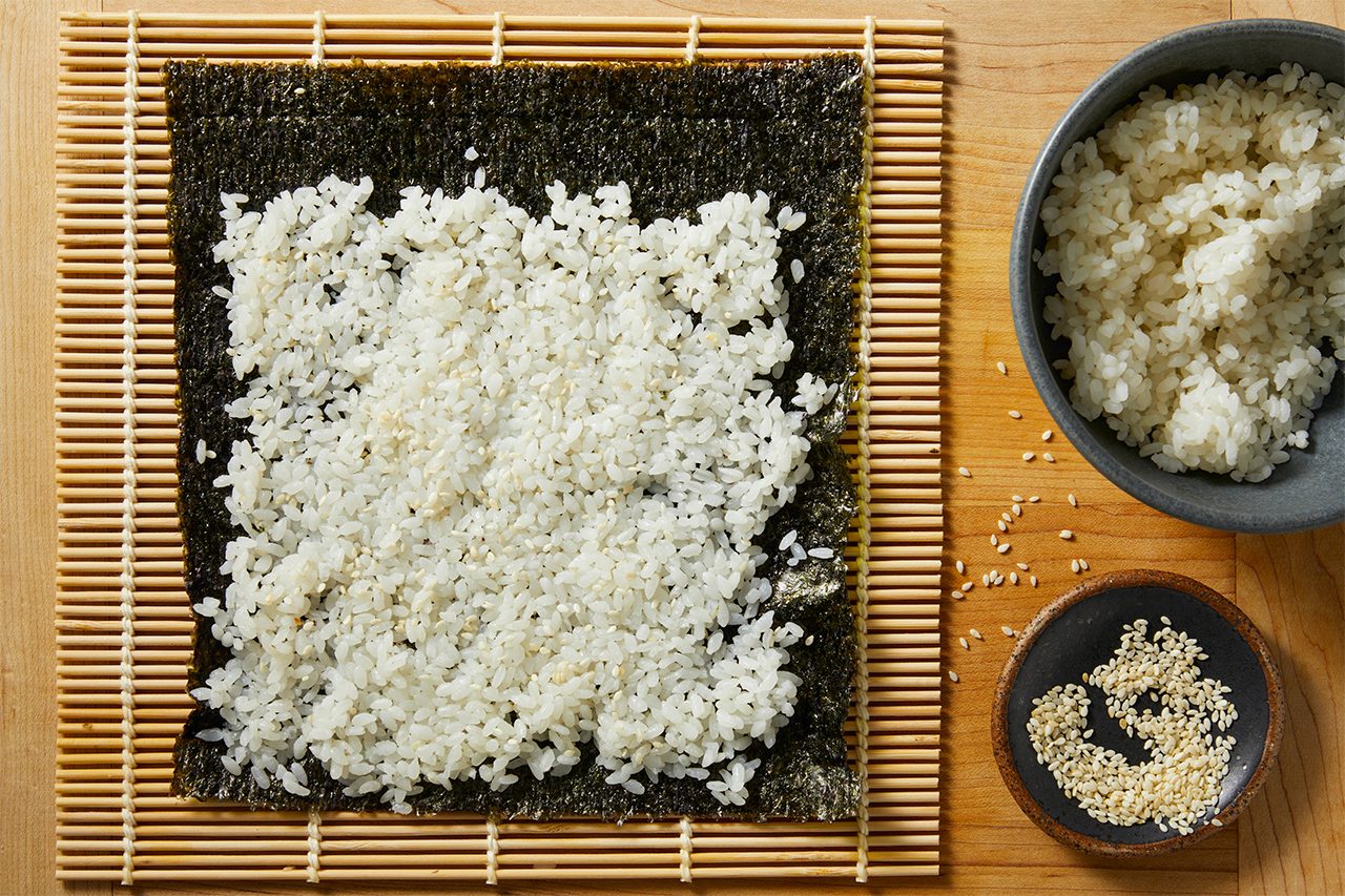 Overhead horizontal image of sushi rice being spread evenly onto a sheet of nori on a bamboo mat. A bowl of cooked sushi rice and a small dish of sesame seeds are visible to the right.