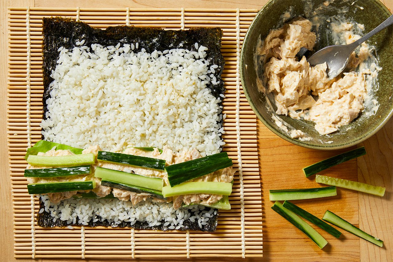 Overhead horizontal image of ingredients arranged on a bamboo rolling mat, ready for sushi preparation. A nori sheet is layered with white sushi rice, a creamy tuna mixture, cucumber strips, and lettuce leaves. A bowl of the tuna mixture and extra cucumber pieces are visible on a wooden cutting board next to the mat.